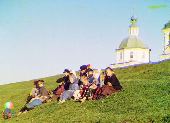 Russian children sit on the side of a hill near a church and bell tower near White Lake, in Russia, 1909